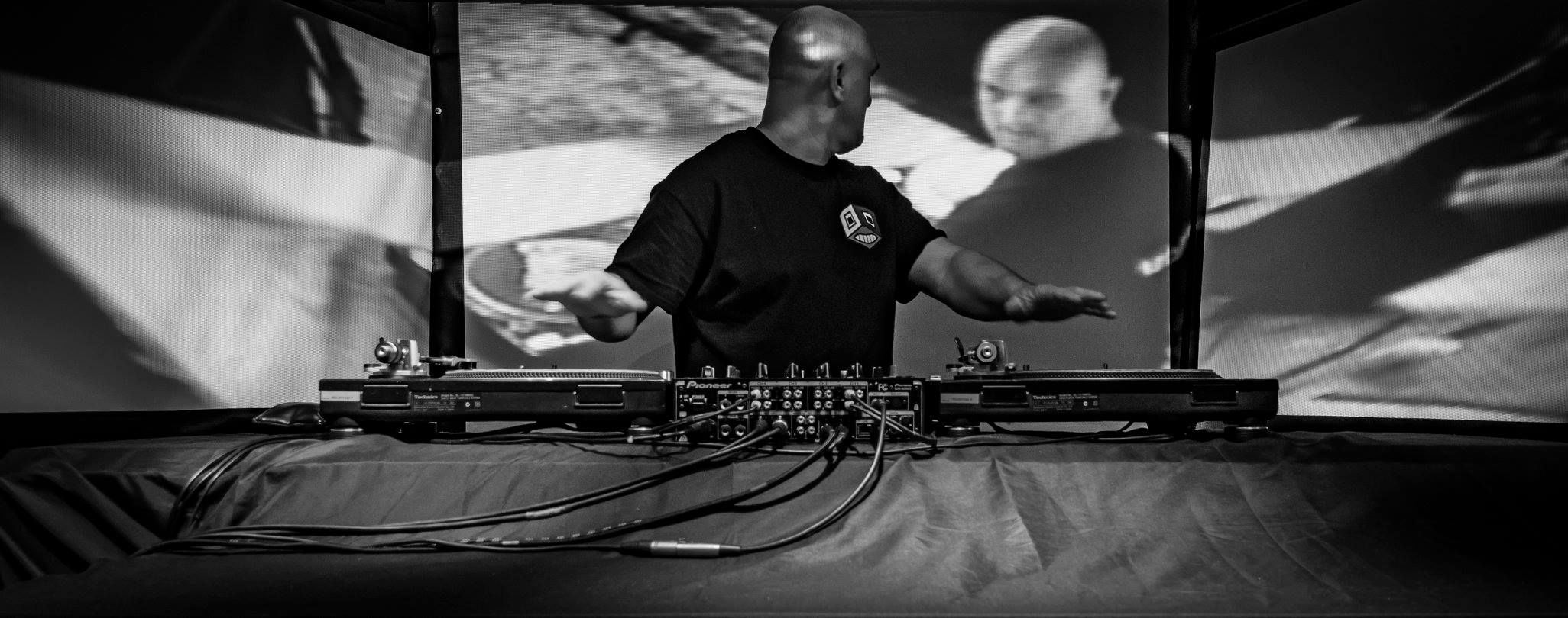 A person standing at a set of turntables, looking over their shoulder at themselves projected on the screen